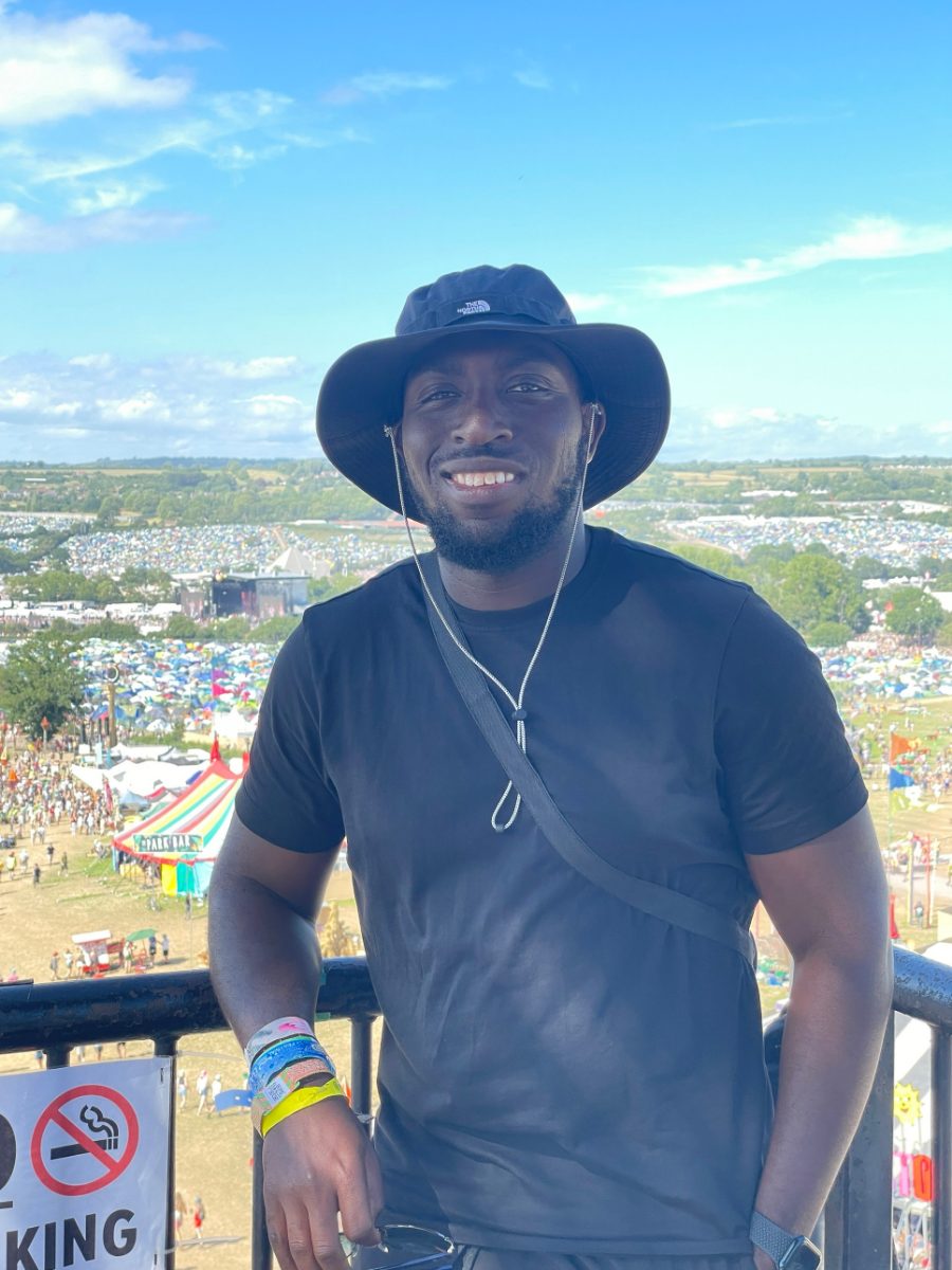 Rasheed smiling on a viewing platform above Glastonbury, with the Pyramid Stage and campsites behind him